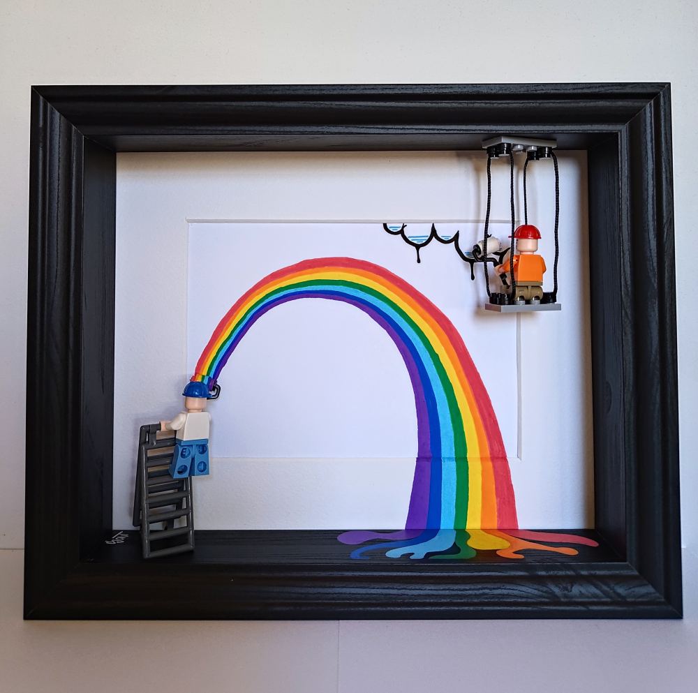 A rainbow painted in a black box frame. One tiny painter on a ladder rolls it on from the left while a second, hanging on a swing stage, paints clouds. The rainbow runs off the paper and pools on the frame's floor.